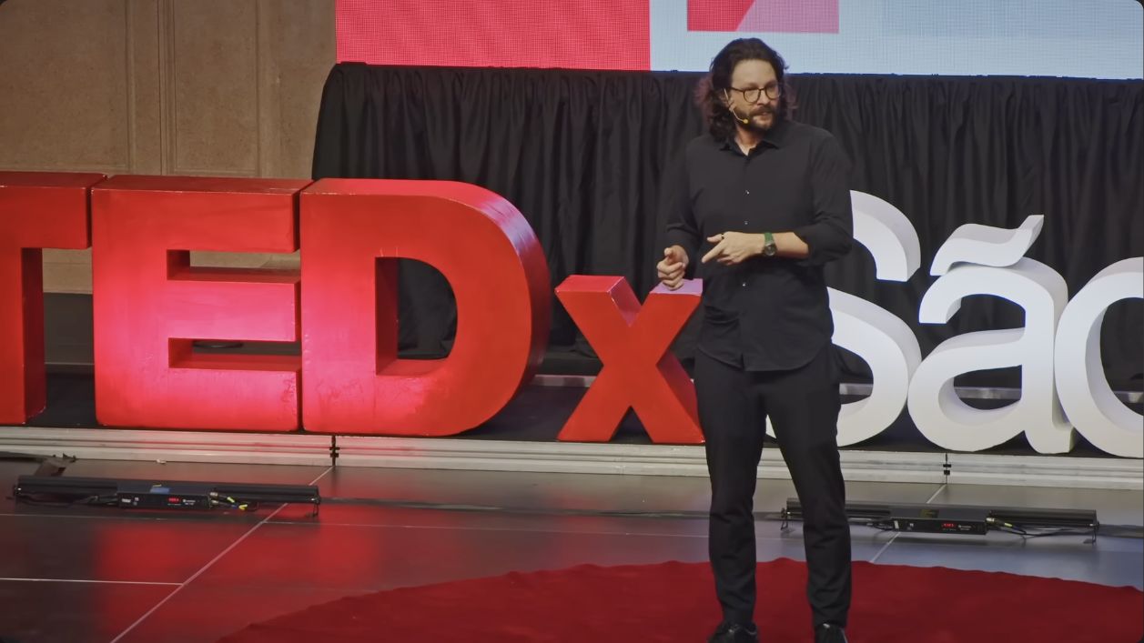Miguel speaking on the TEDxSaoPaulo stage, with the TEDx letters behind him.